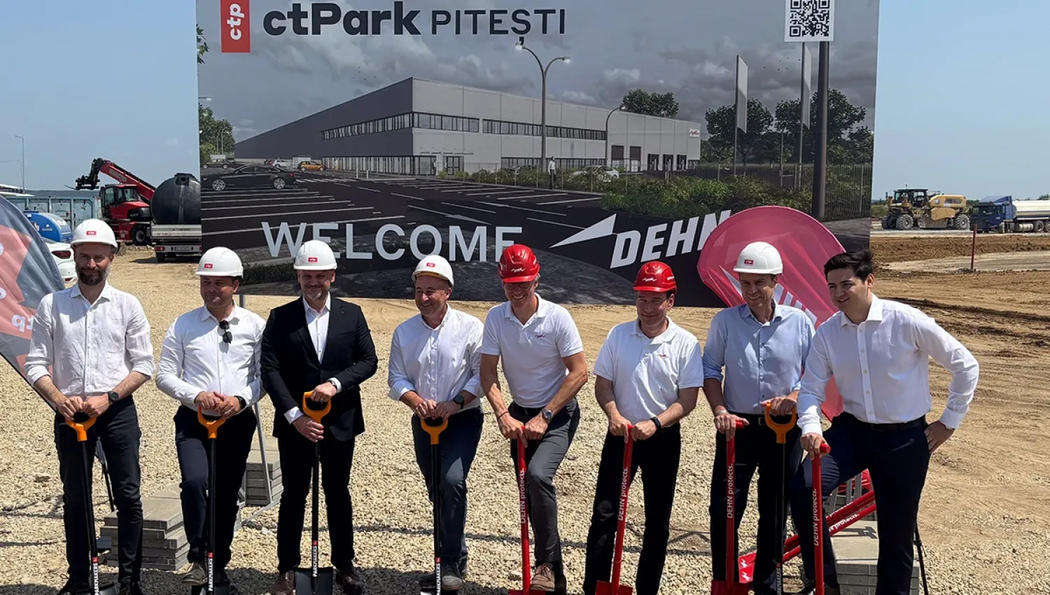 Group of people wearing hard hats holding shovels at a construction site, standing in front of a sign reading “ctPark PITESTI” and “WELCOME DEHN”.