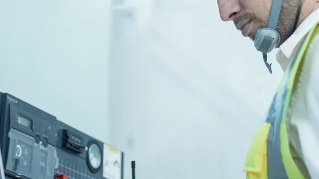 A man wearing a safety helmet and high-visibility waistcoat is standing at an electrical installation with a laptop.