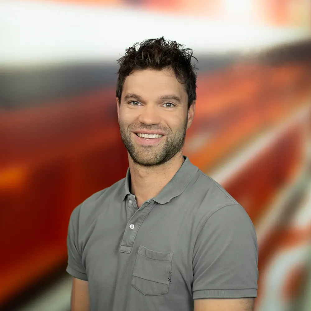 Smiling man with short dark hair and light stubble, wearing a gray polo shirt, standing in front of a blurred red and orange background.