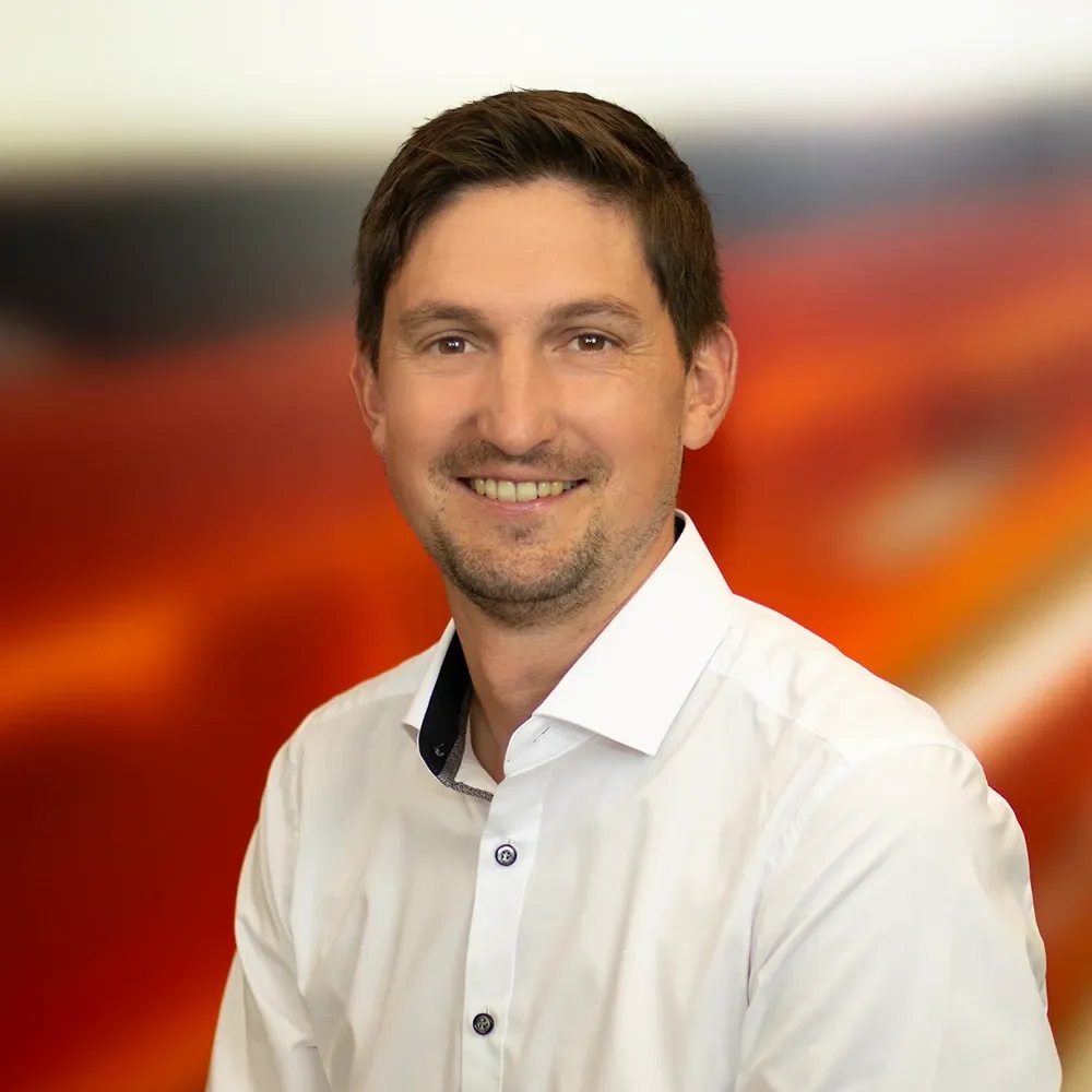 Smiling man wearing a white button-up shirt, standing in front of a blurred red and orange background.